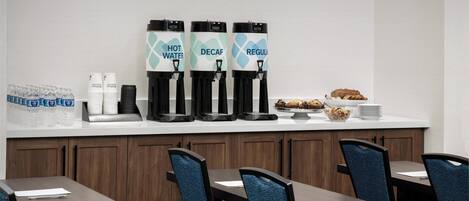 Una colazione con dispenser di acqua calda, caffè decaffeinato e caffè normale, una pila di tazze bianche e una ciotola di muffin.