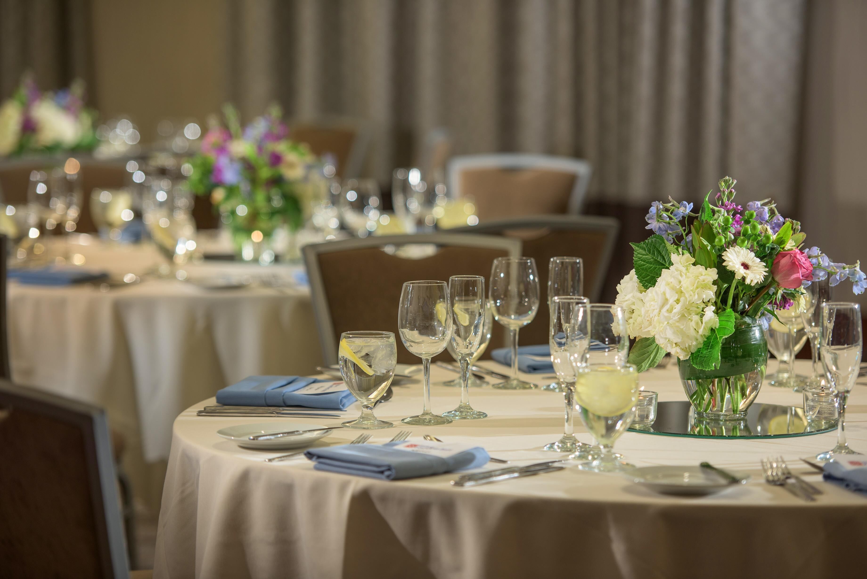 A dining area with round tables set for a meal, each with a vase of flowers and clear glassware.