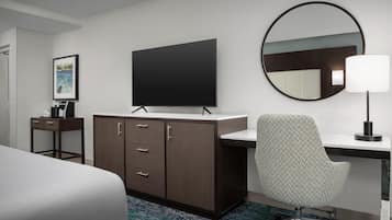 Habitación de hotel con cama, escritorio con silla, televisor de pantalla plana, espejo y un cuadro en la pared.