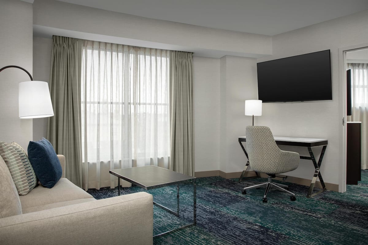 Ein Hotelzimmer mit einer Couch, einem Schreibtisch, einem Stuhl, einem Fernseher und einem großen Fenster mit Vorhängen.
