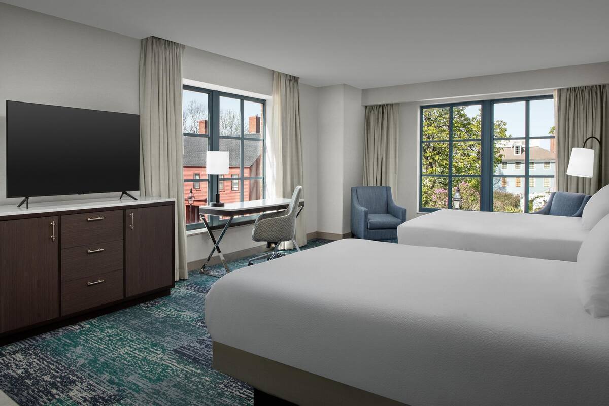 Ein Hotelzimmer mit zwei Betten, einem Schreibtisch, einem Fernseher und großen Fenstern mit Vorhängen.
