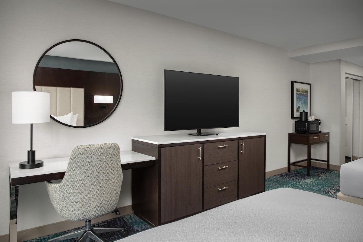 Ein Hotelzimmer mit einem Bett, einem Schreibtisch mit Stuhl, einem Fernseher, einem Spiegel, einem kleinen Tisch mit einer Mikrowelle und einem Bild an der Wand.