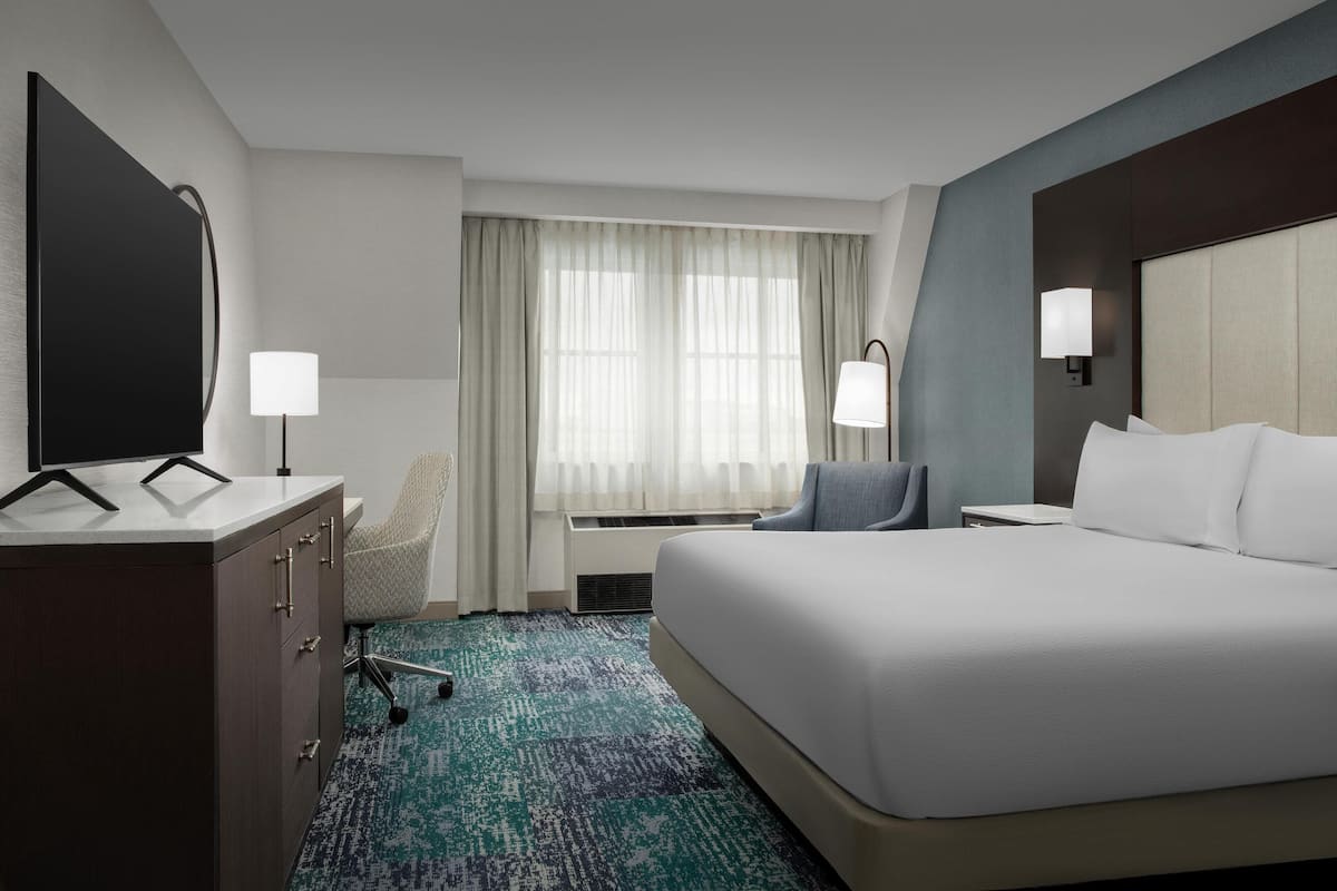 Ein Hotelzimmer mit einem großen Bett, einem Schreibtisch mit Flachbildfernseher, einem Sessel, zwei Nachttischlampen und einem Fenster mit durchsichtigen Vorhängen.