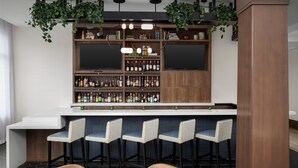 A bar area with a counter, multiple shelves of bottles, hanging plants, and bar stools. - Hilton Garden Inn Portsmouth Downtown (Portsmouth)