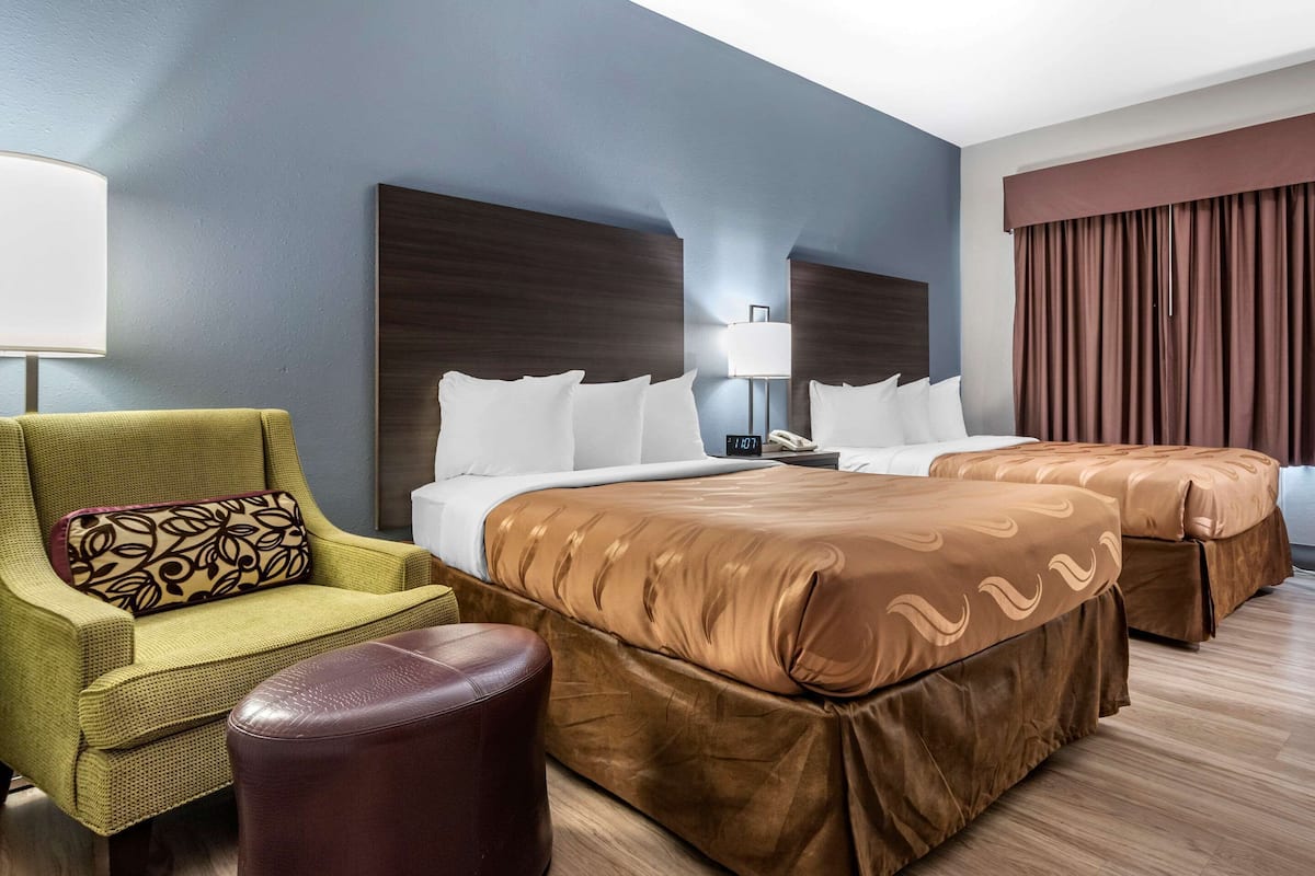 A hotel room with two beds, a green armchair, a brown ottoman, and a large window with curtains.