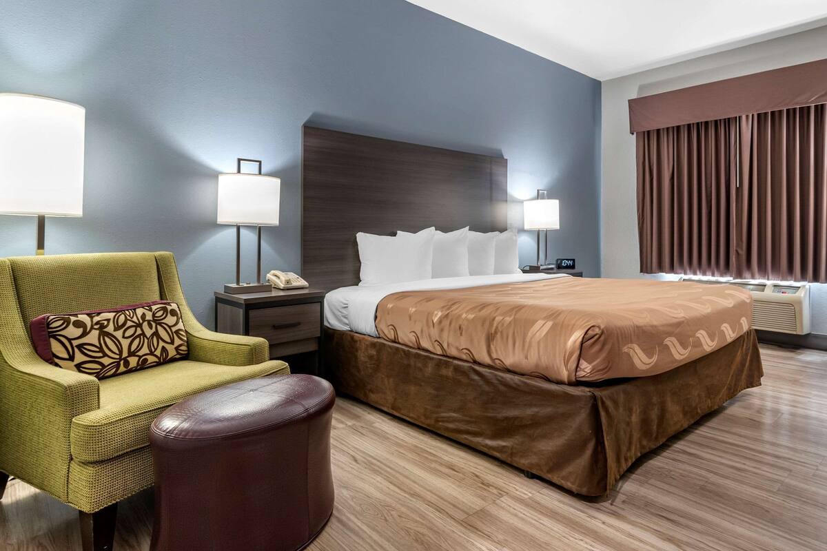 A hotel room with a large bed, a green armchair, a brown ottoman, two bedside lamps, and a nightstand with a phone.