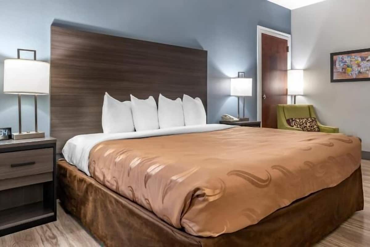 A hotel room with a bed, bedside lamps, a chair, a nightstand, and a painting on the wall.