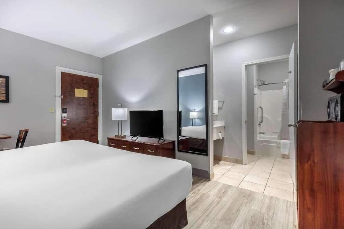 A hotel room with a bed, a television, a desk with drawers, a mirror, and a bathroom visible through an open door.
