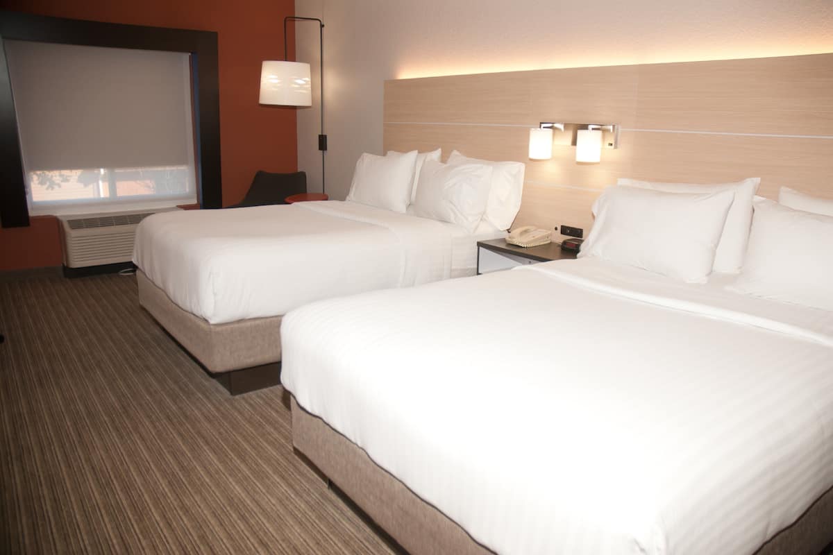 A hotel room with two beds, a bedside table with a phone, a lamp, and an air conditioning unit.