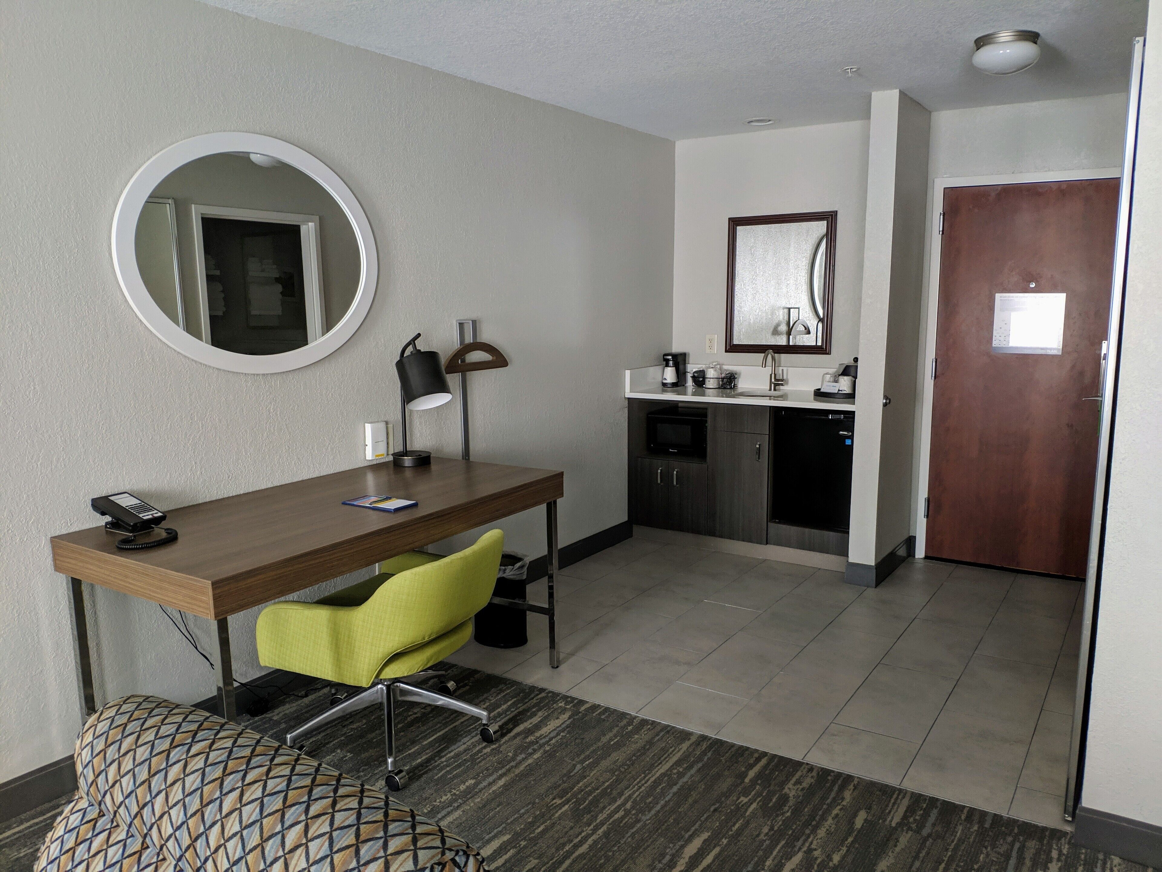 A compact hotel room with a wooden desk, a green chair, a round mirror, a small kitchenette, and a door.