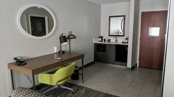 A compact hotel room with a wooden desk, a green chair, a round mirror, a small kitchenette, and a door.