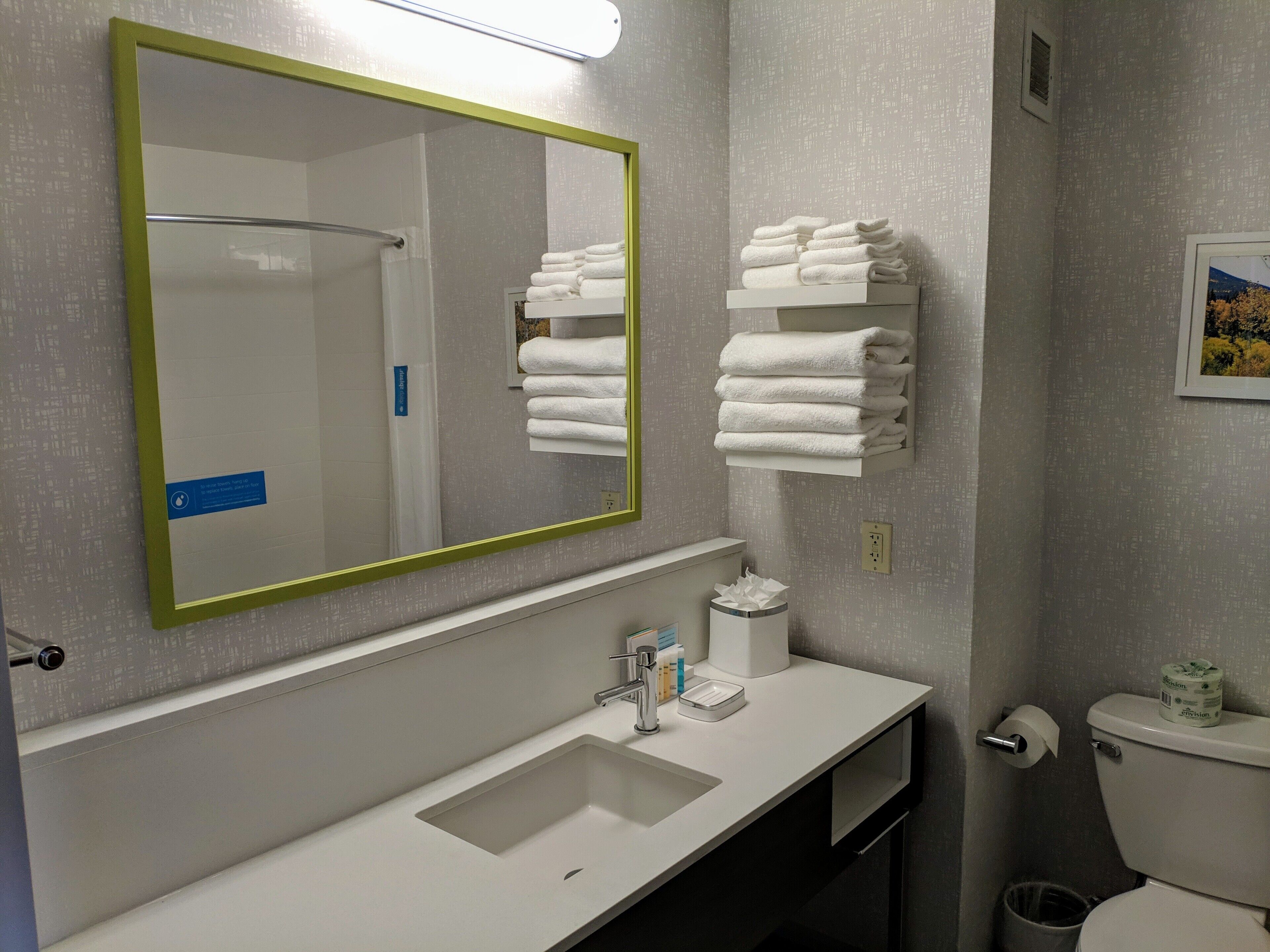 A bathroom with a large mirror, a sink, a toilet, and a stack of white towels.