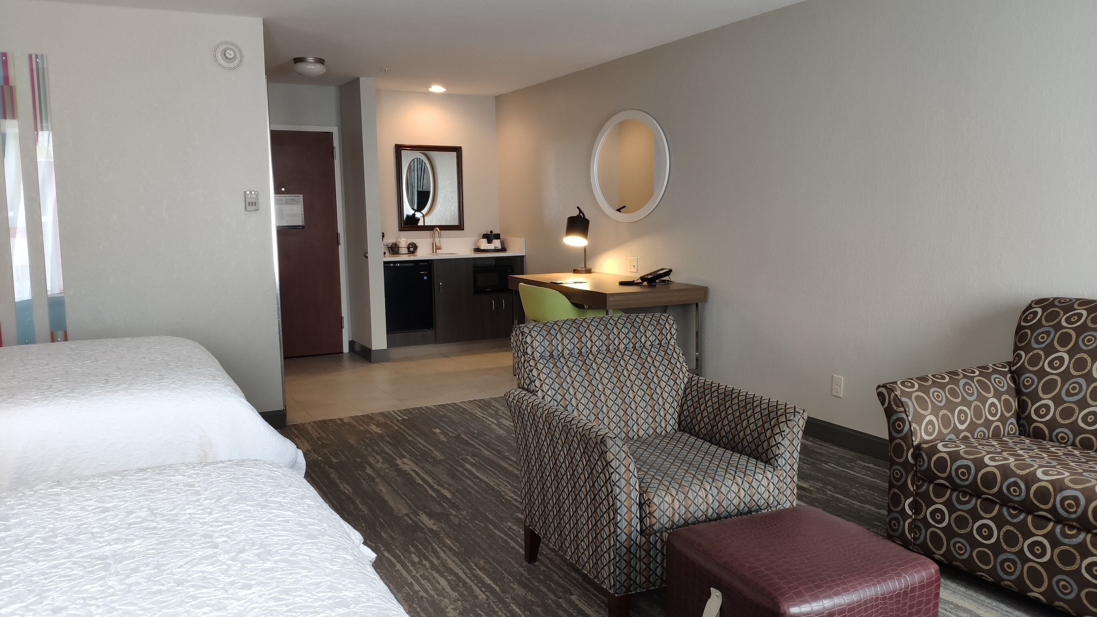 Ein Hotelzimmer mit zwei Betten, einem Schreibtisch mit Spiegel, einem gemusterten Sessel und einem kleinen Hocker.