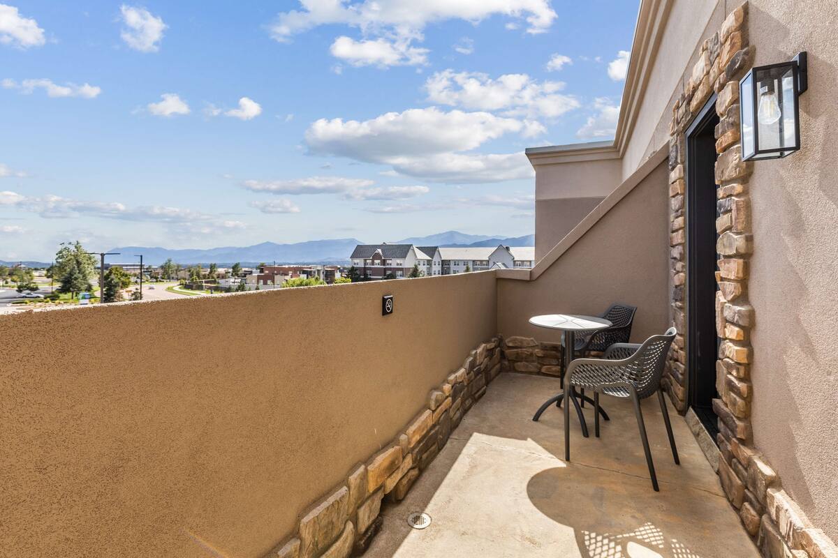 A balcony with a stone wall, a table, and two chairs, overlooking a residential area and mountains.