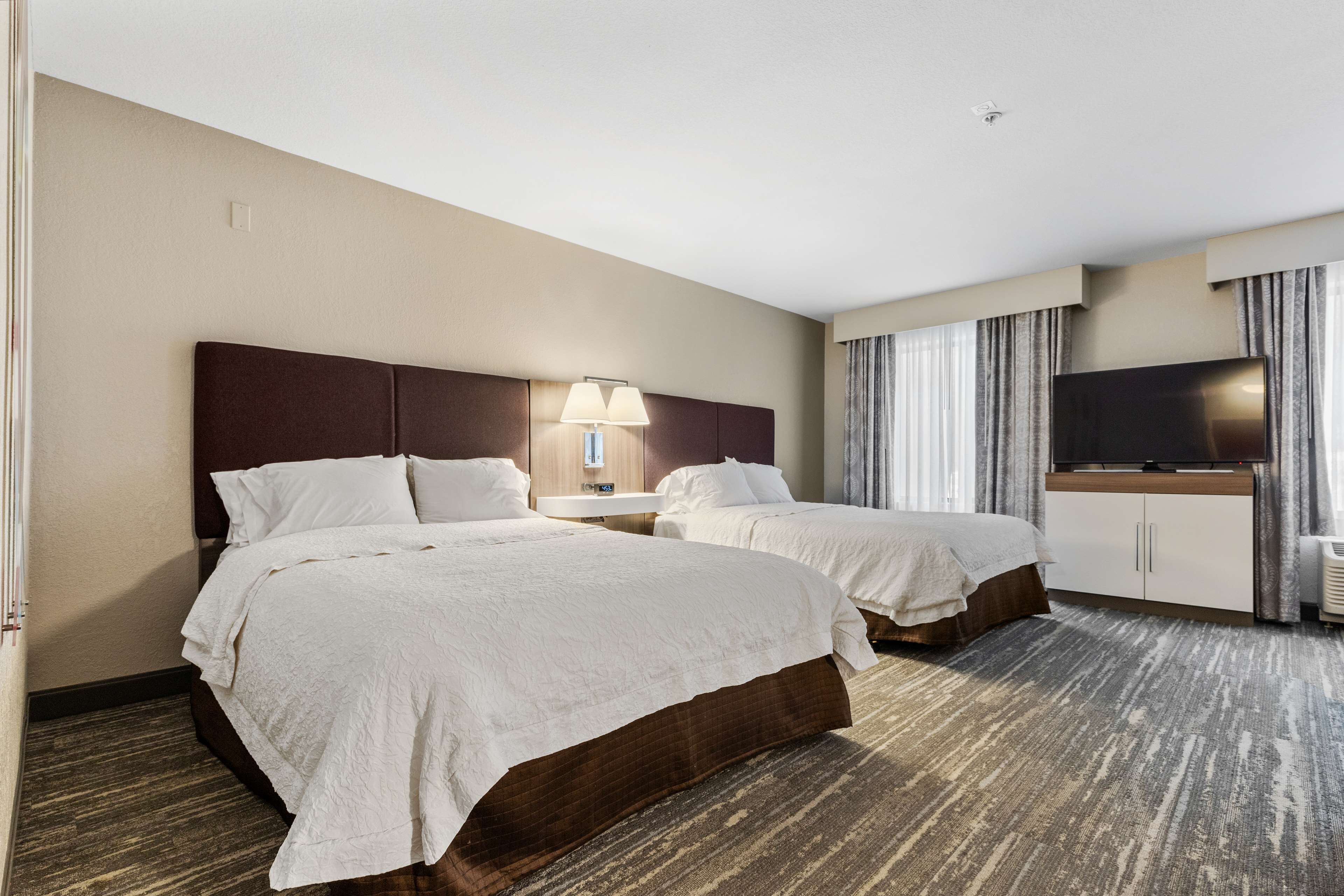 Habitación de hotel con dos camas, televisión, lámpara y un amplio ventanal con cortinas.