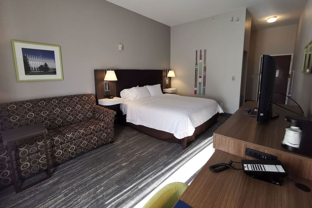 A hotel room with a bed, two bedside lamps, a TV, a desk with a computer, a chair, and a patterned sofa.