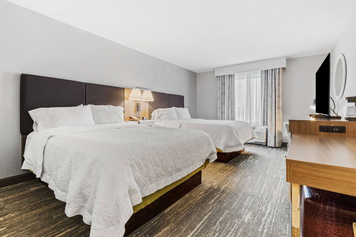 A hotel room with two beds, a desk, a chair, a TV, and a window with curtains.