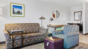A modern hotel room with a patterned sofa, a patterned armchair, a small table, a purple ottoman, a round mirror, a desk, and a kitchenette.