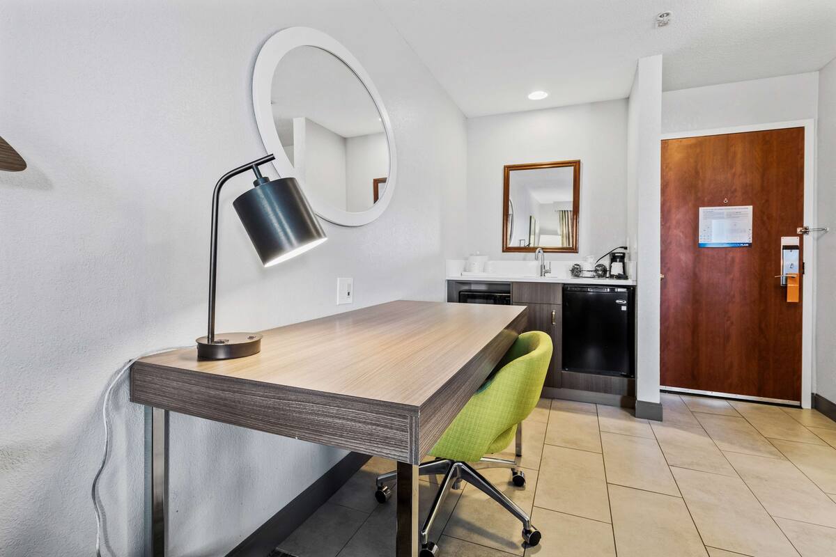 A modern hotel room with a wooden desk, a green office chair, a round mirror, a sink, and a wooden door.