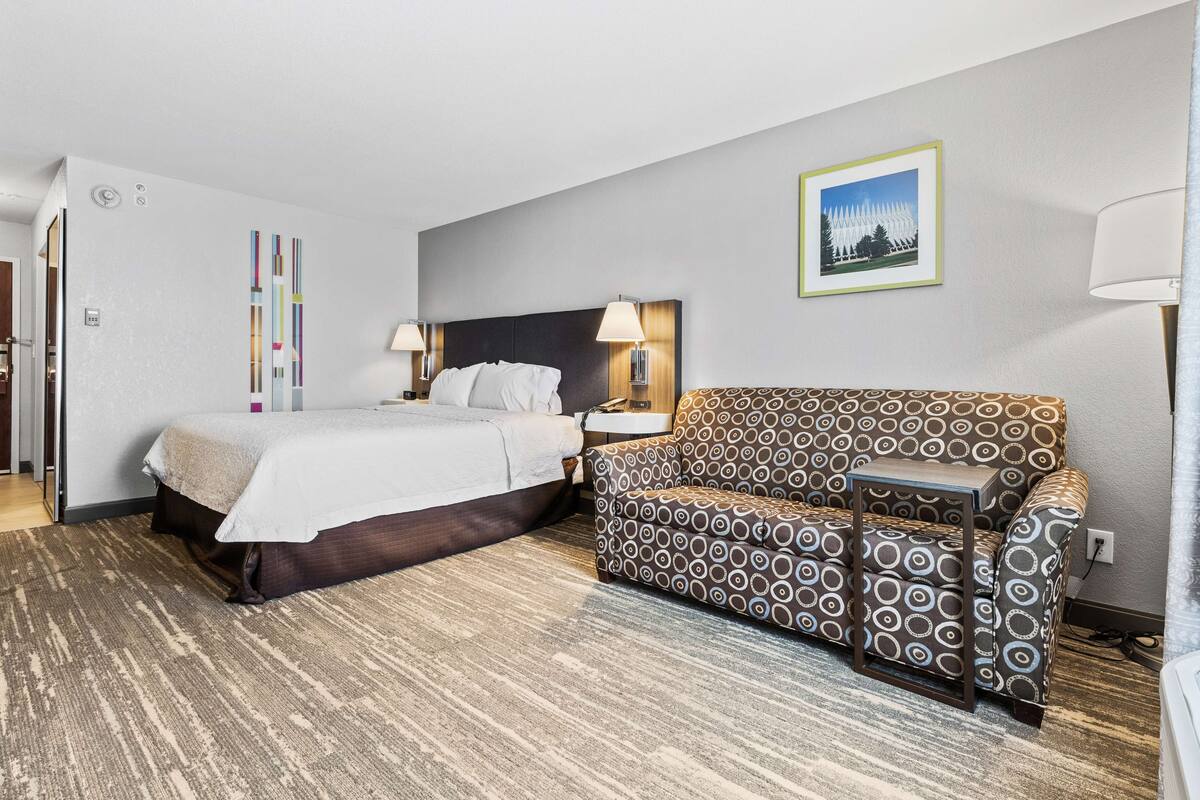 A hotel room with a bed, a patterned sofa, a nightstand, and a wall-mounted lamp.