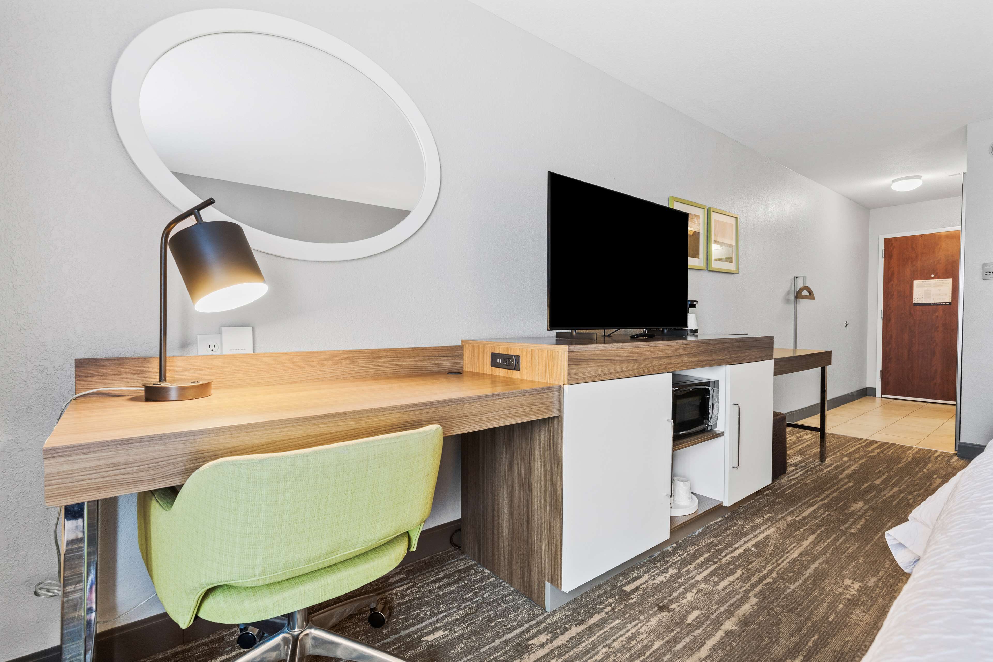 A hotel room with a wooden desk, a green chair, a flat-screen TV, a round mirror, and a small kitchenette.