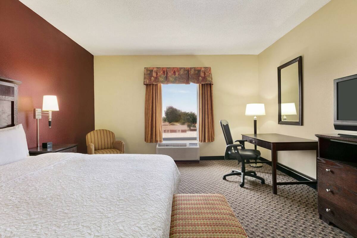 A hotel room with a bed, a desk, a chair, a dresser, a TV, and a window with curtains.