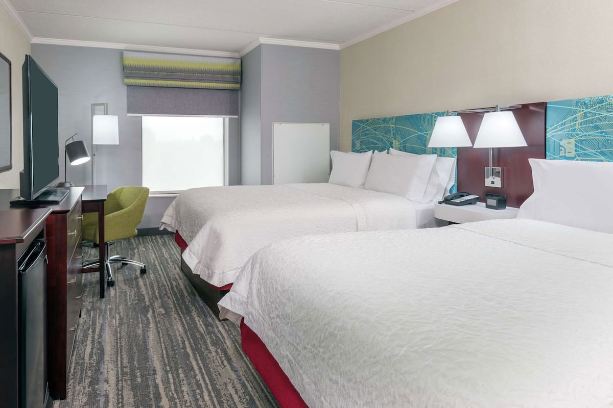 A hotel room with two beds, a desk with a chair, a lamp, and a painting on the wall.