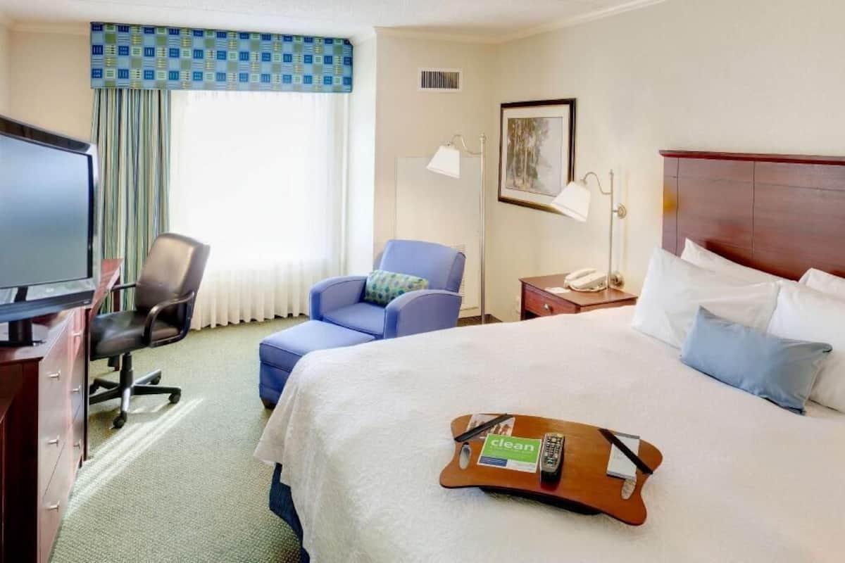 A hotel room with a bed, a desk with a TV, a chair, a bedside table with a lamp, and a window with curtains.