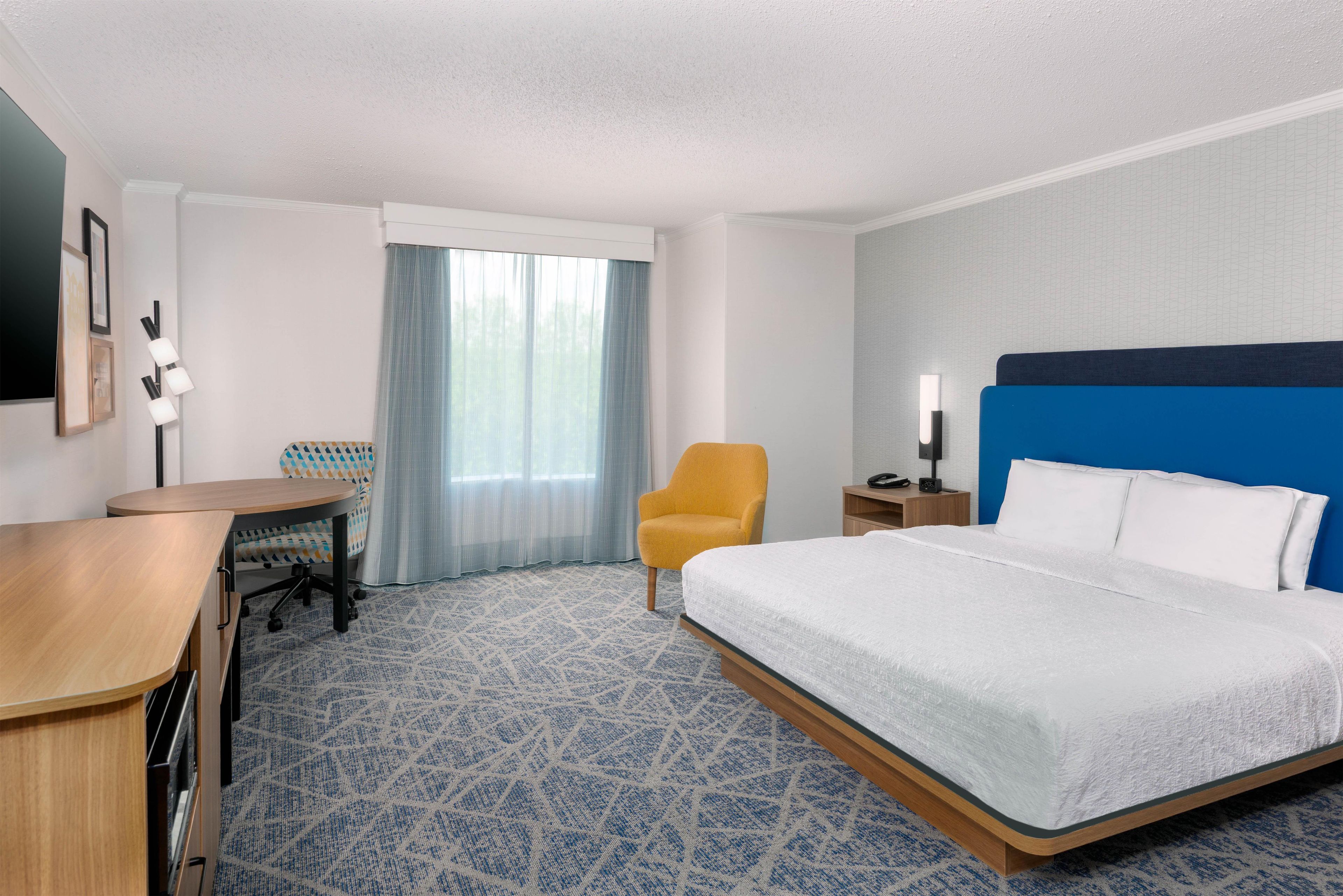 A hotel room with a bed, a desk, a chair, a nightstand, and a window with curtains.