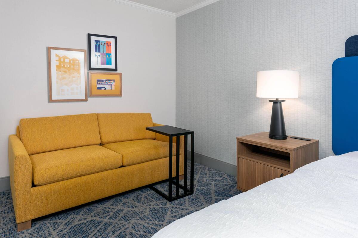 A hotel room with a sofa, a small table with a lamp, a bed, and two framed pictures on the wall.