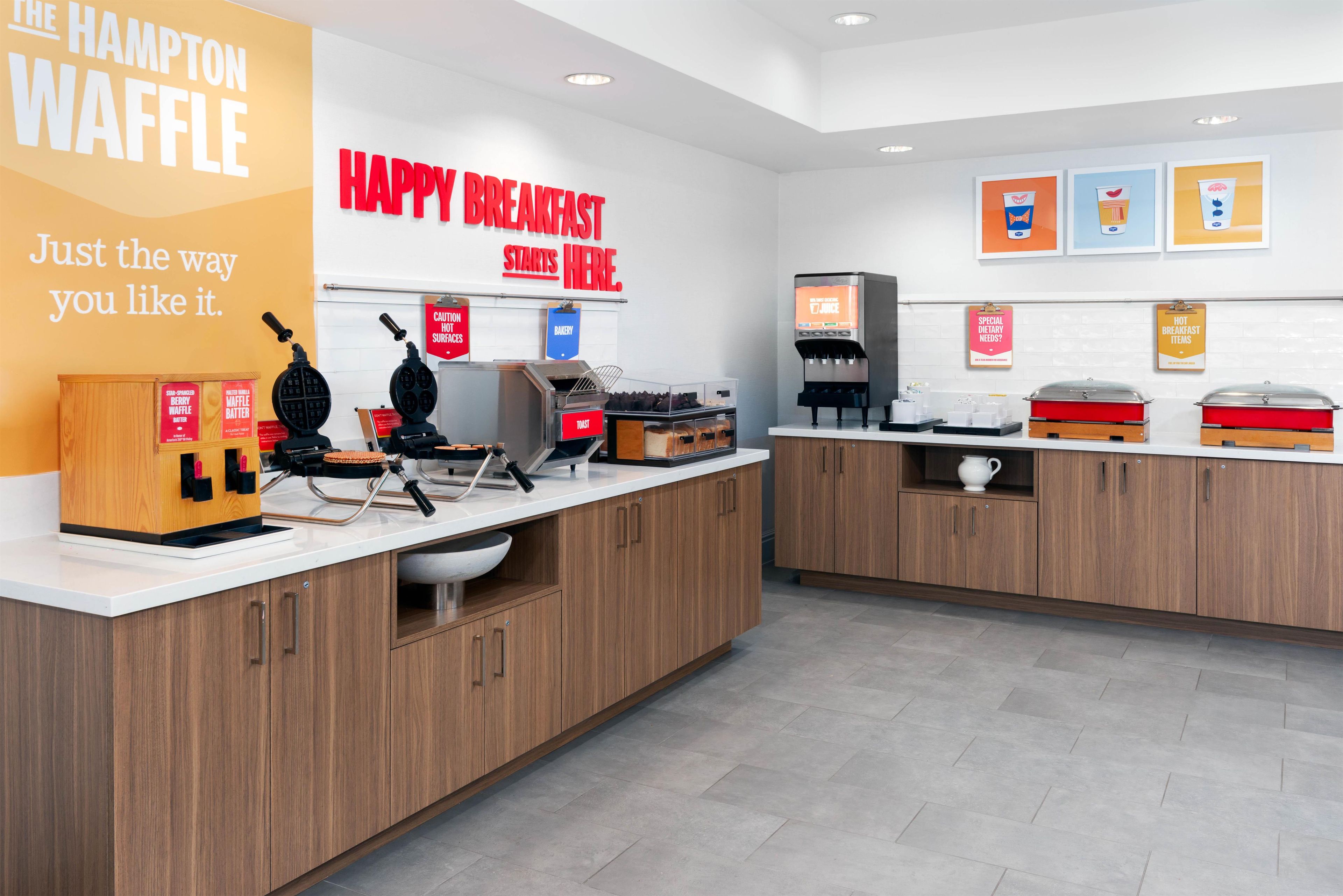 A clean, modern breakfast area with waffle makers, a coffee station, and a sign reading 'HAPPY BREAKFAST STARTS HERE.'