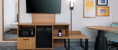 A hotel room with a flat-screen TV, a microwave, a mini-fridge, a coffee maker, a wooden desk, a round table, and a patterned carpet.