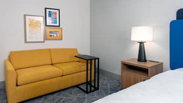 Habitación de hotel con sofá, una mesita de noche con lámpara, cama y dos cuadros en la pared.