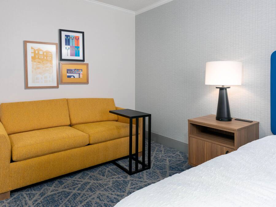 Habitación de hotel con sofá, una mesita de noche con lámpara, cama y dos cuadros en la pared.