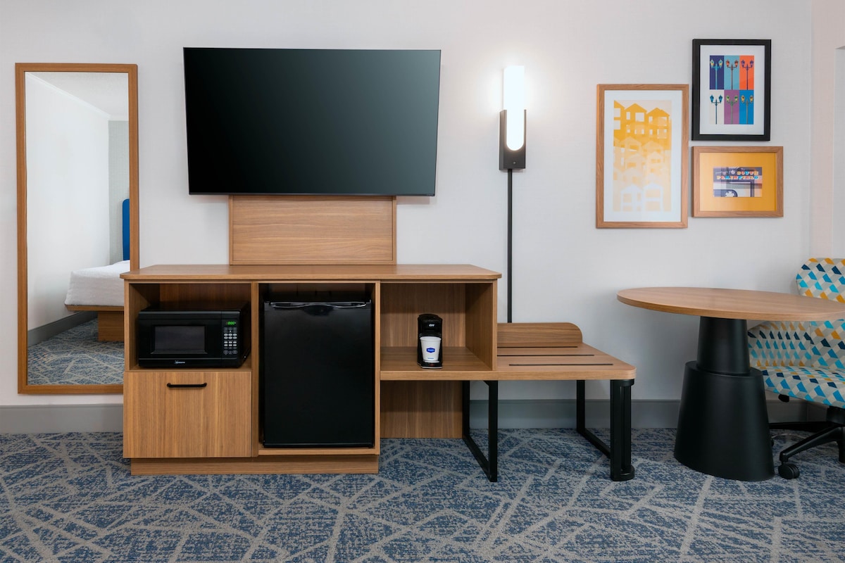 A hotel room with a flat-screen TV, a microwave, a mini-fridge, a small table, a chair, and a wall-mounted light.