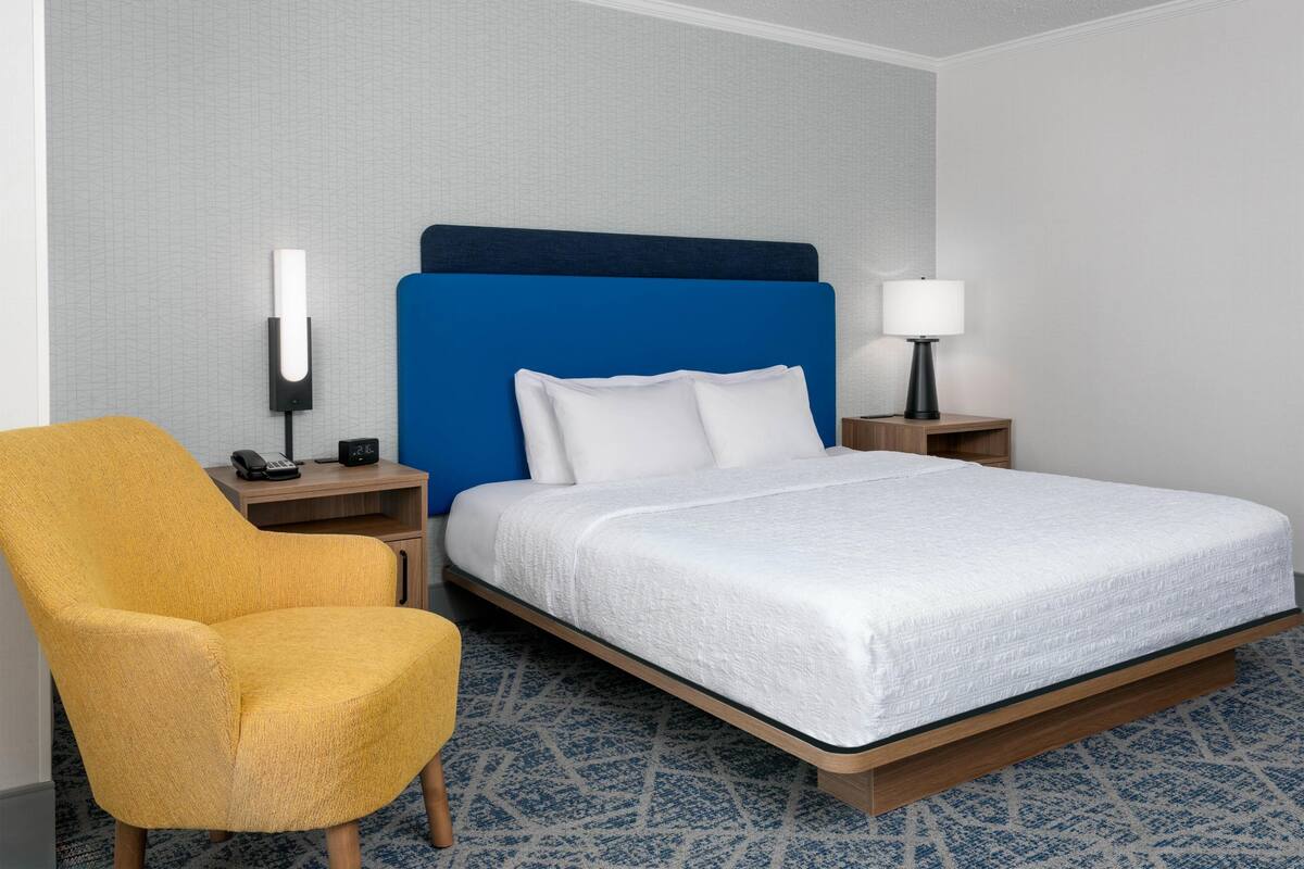 A hotel room with a bed, a chair, a bedside table, a lamp, and a wall-mounted phone.
