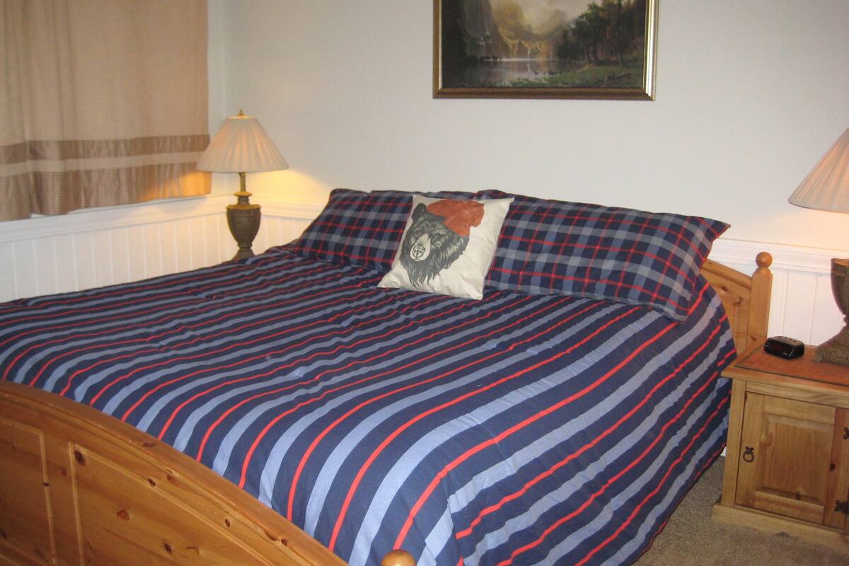 A neatly made bed with striped bedding, a wooden headboard, a bedside table with a lamp, and a framed painting on the wall.