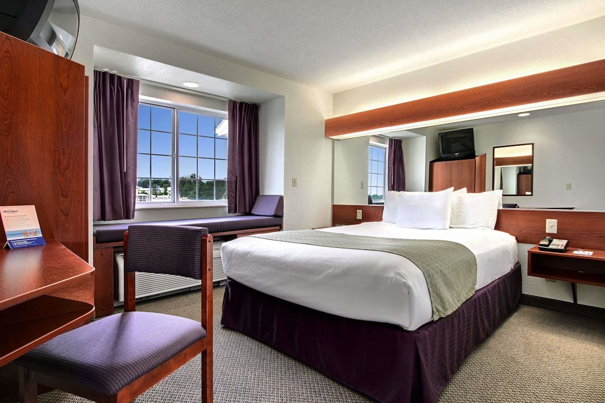 A hotel room with a large bed, a desk, a chair, a window with curtains, a mirror, and a television on a wooden cabinet.