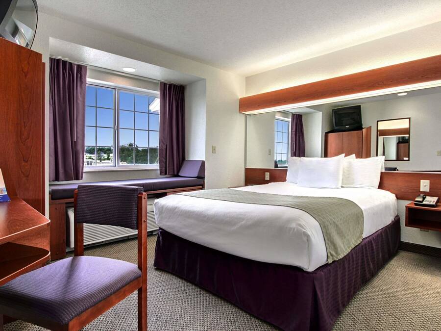 Habitación de hotel con una cama grande, un escritorio, una silla, un ventanal con cortinas, un espejo y un televisor sobre un mueble de madera.
