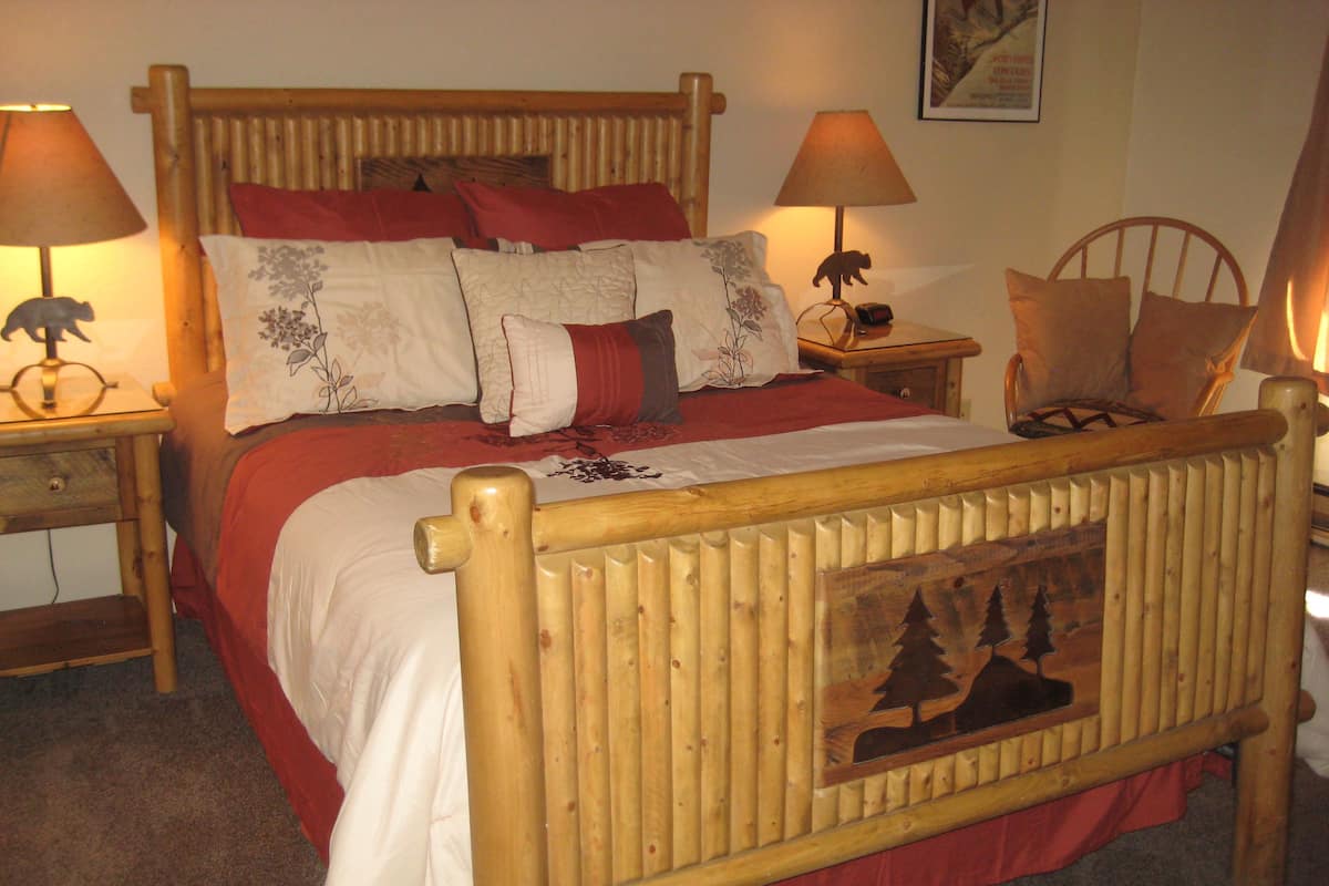 Uma cama de madeira com cabeceira retratando uma cena de floresta, ladeada por duas mesas de cabeceira com abajures, e um cobreleite vermelho.