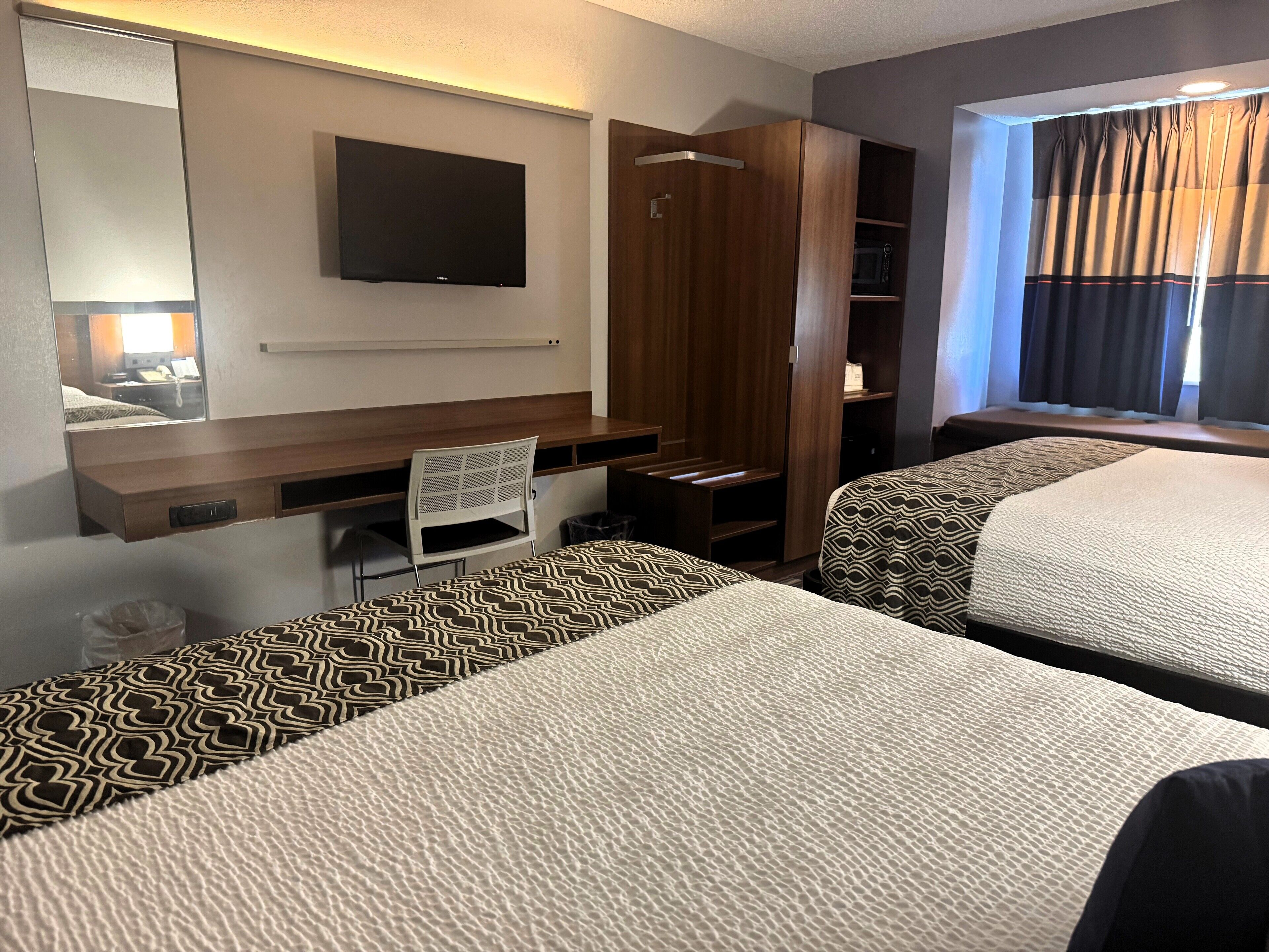 Habitación de hotel con dos camas, un escritorio, una silla, un armario, televisión y ventana con cortinas.