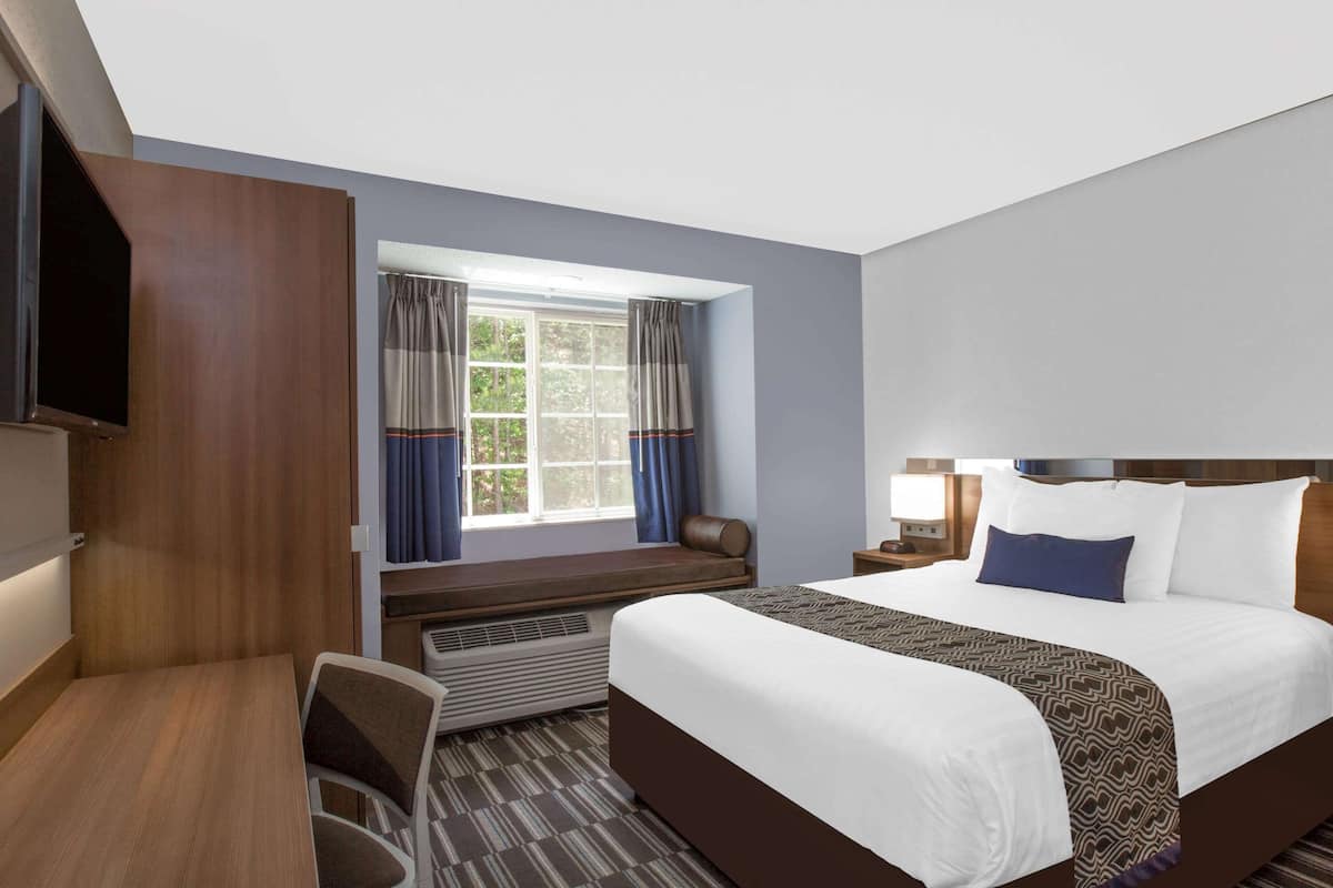 A hotel room with a large bed, a desk with a chair, a TV mounted on the wall, and a window with curtains.