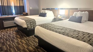 Habitación de hotel con dos camas, una ventana con cortinas, una mesita de noche con lámpara y suelo alfombrado.