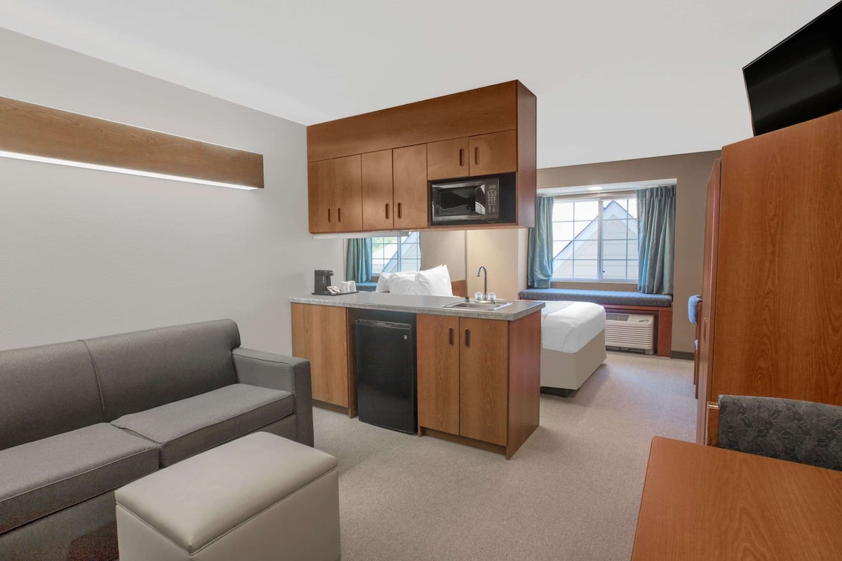 A compact hotel room with a kitchenette, a sofa, a small dining table, and a bed with a window view.