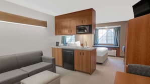 Habitación de hotel compacta con kitchenette, sofá, pequeña mesa de comedor y cama con vista al exterior.