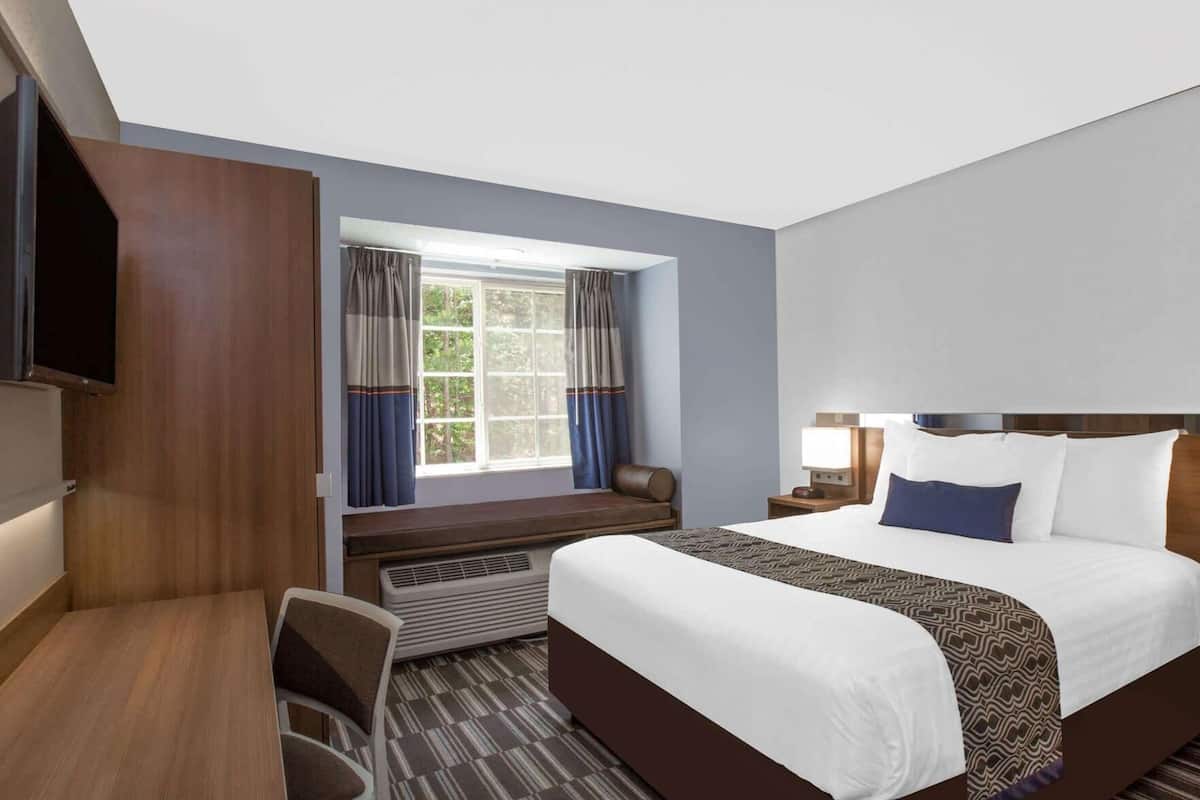 A hotel room with a bed, a desk, a chair, a TV, a window with curtains, and a heater.