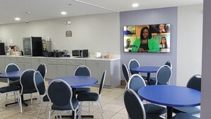 Una sala de conferencias con una pantalla grande mostrando una videoconferencia, varias mesas y sillas, y una zona de kitchenette al fondo.