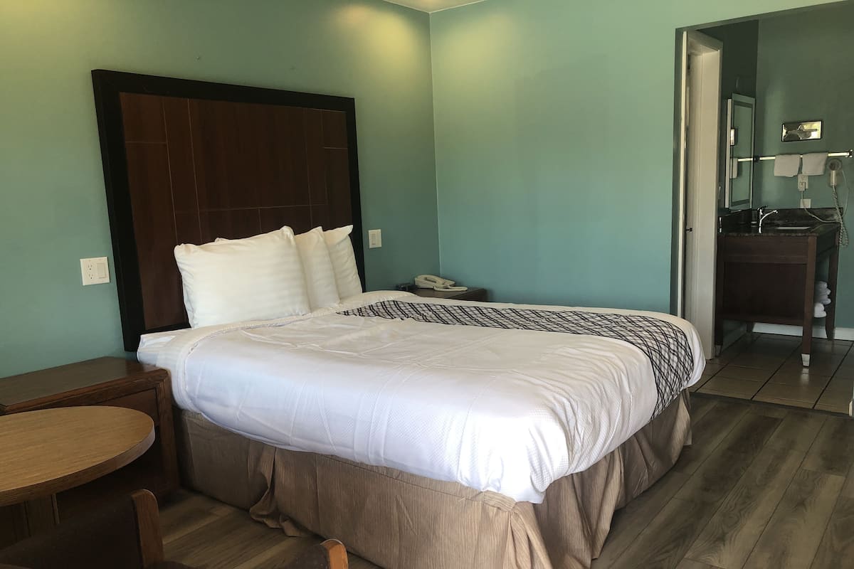 Una cama bien tendida con cabecera, una mesita de noche y una silla. Se ve un baño a través de una puerta abierta.