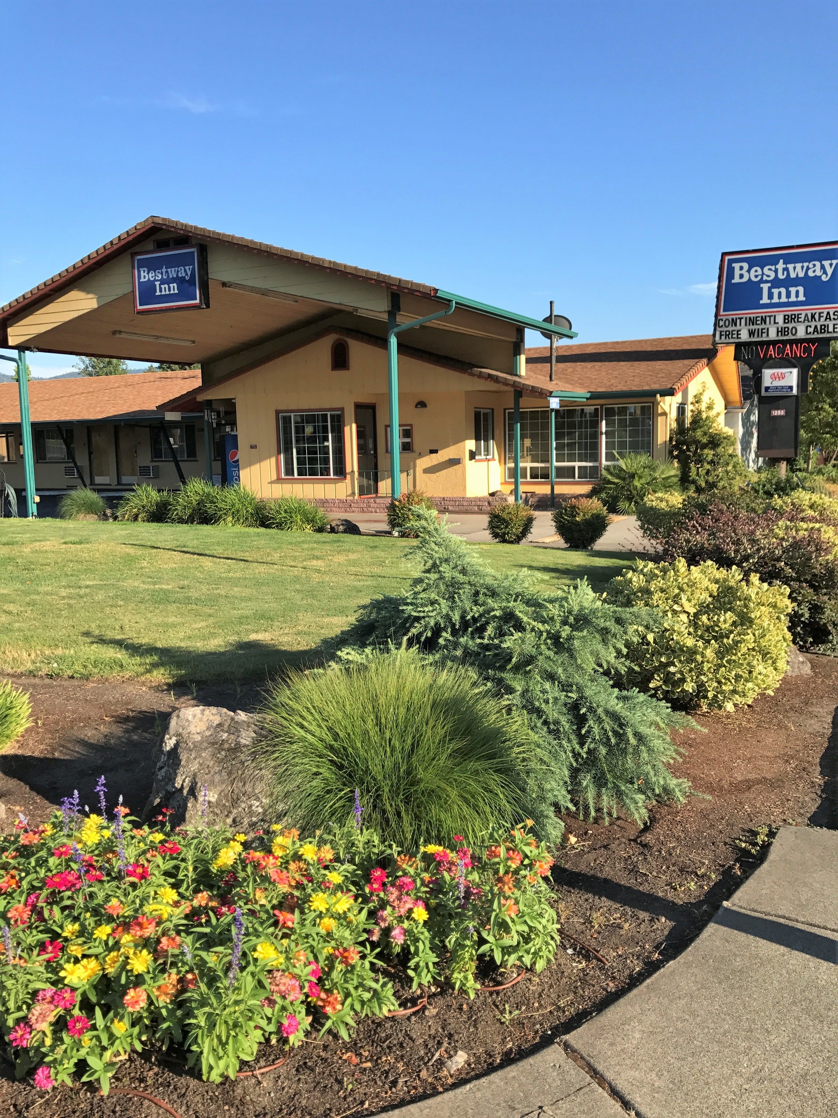 A Bestway Inn with a covered entrance, a sign indicating no vacancy, and a landscaped garden with colorful flowers and shrubs.