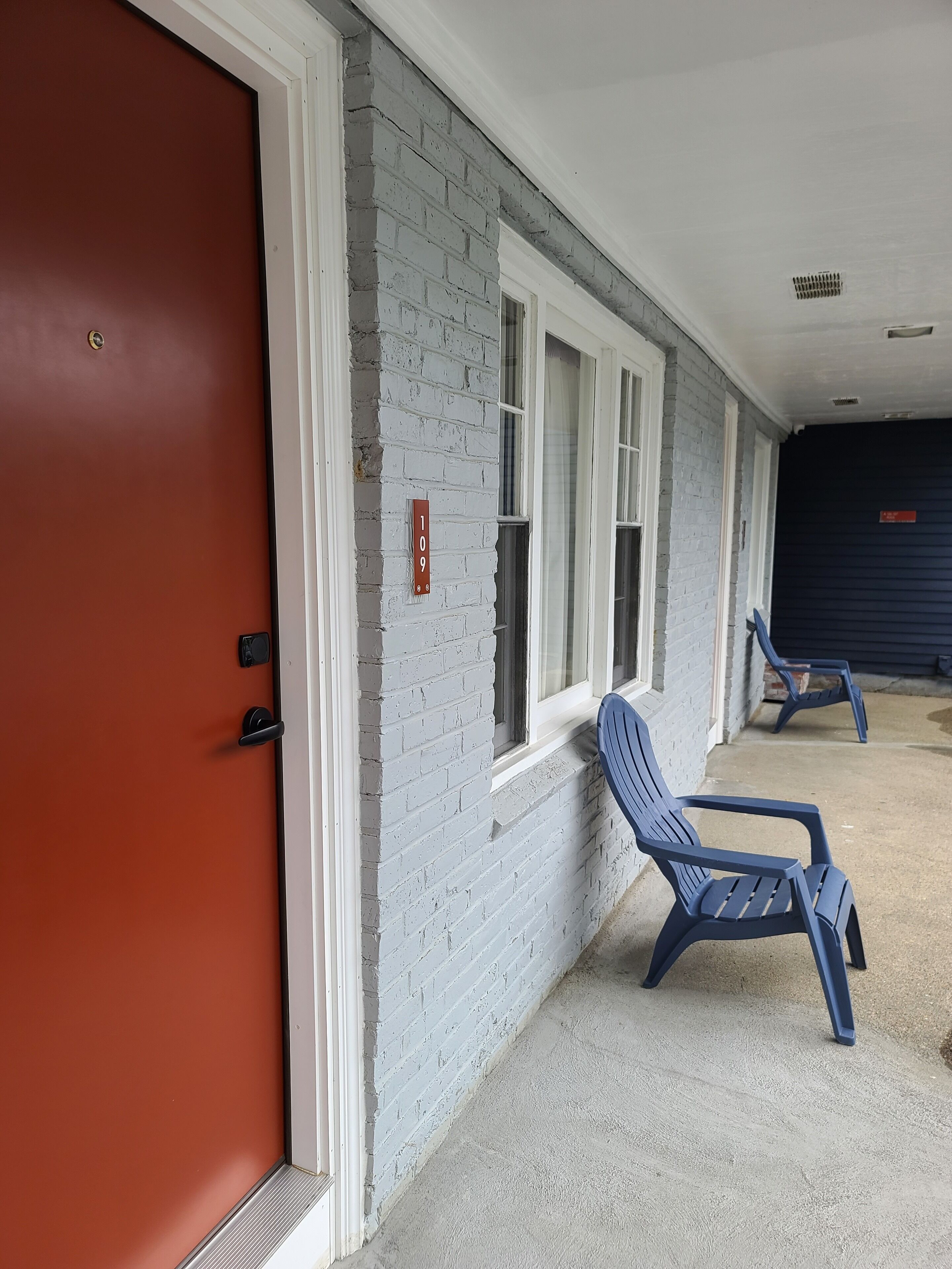 Fachada de edificio con una puerta roja, una silla azul y un letrero con número en la pared.